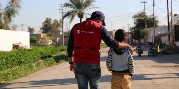 operatore save the children con maglietta rossa cammina al fianco di un bambino, entrambi li vediamo di spalle 
