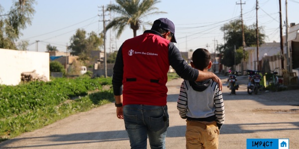 operatore save the children con maglietta rossa cammina al fianco di un bambino, entrambi li vediamo di spalle 
