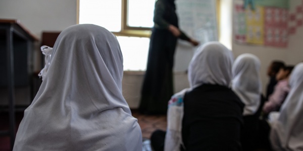 ragazze di spalle, sedute per terra in un aula scolastica, indossano un velo bianco e guardano verso la maestra