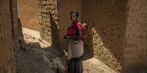 Donna ruandese in piedi tra due mura porta sulle spalle un bambino neonato nella fascia. Lei indossa una maglia rosa e una lunga gonna marrone.