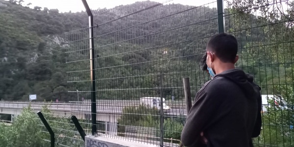 Ragazzo preso di spalle indossa una felpa grigia e guarda davanti a sé. Si trova all aperto e di fronte a sé ha una rete e oltre una collina alberata.