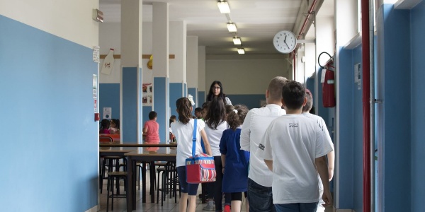 Bambini in fila nel corridoio di una scuola sono inquadrati di spalle mentre camminano verso l insegnate sullo sfondo girata verso di loro