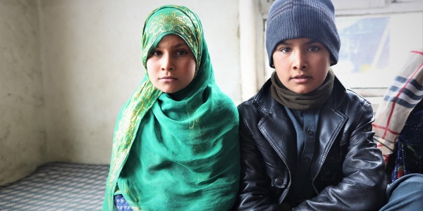 due bambini afghani seduti a terra, un maschio e una femmina. Lei indossa un chador verde e lui un berretto blu e una giacca di pelle nera..