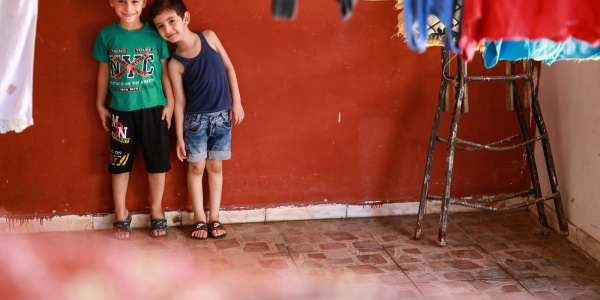 due bambini ripresi in lontananza all interno di una stanza con le pareti rosse sono appoggiati al muro uno a fianco dell altro. davanti a loro i panni stesi.