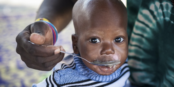 Primo piano di un bambino del Burkina Faso che viene imboccato con un cucchiao da una donna di cui si vede solo la mano e un pezzo di vestito verde