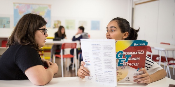 due ragazze sedute al banco di scuola una di loro con un libro di esercizi in mano parlano tra loro