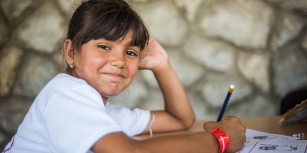 bambina di carnagione chiara-olivastra, con capelli castani legati con frangia, è seduta a un banco di scuola e guarda in camera mentre tiene una matita in mano.