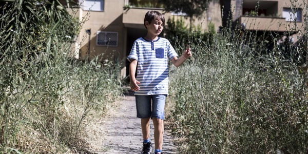 Un bambino con maglietta a righe e pantaloncini cammina in una viale sterrato con ai bordi erba alta