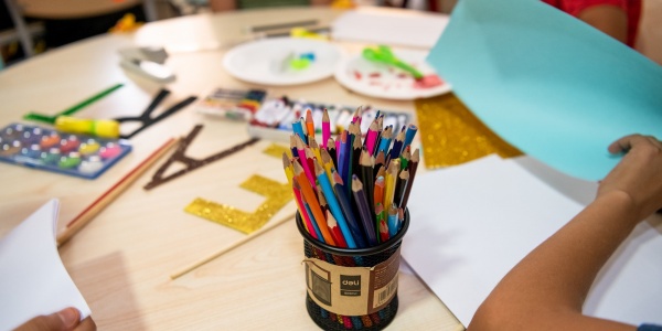 tavolo rotondo con attorno 4 bambini non riconoscibili poiché inquadrati dalle spalle in giù. Sul tavolo materiale da disegno e in primo piano un porta pastelli pieno di pastelli colorati.