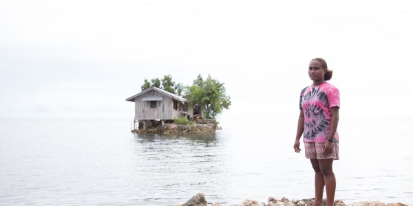 Una ragazza delle Solomon Island è in piedi sulla riva di un lago e la sua casa è un isolotto artificiale alle sue spalle