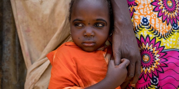 Primo piano di una bambina nigeriana con indosso una maglietta arancione che tiene per mano la mamma