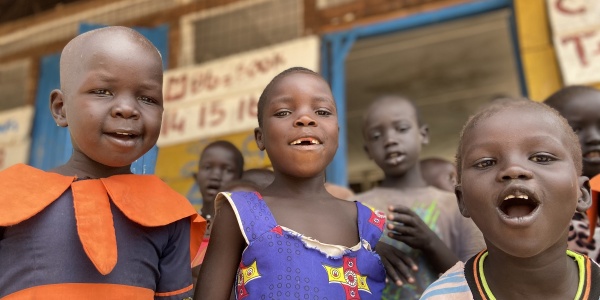 Tre bambini ugandesi in primo piano sorridono in camera. Sullo sfondo struttura in legno della scuola