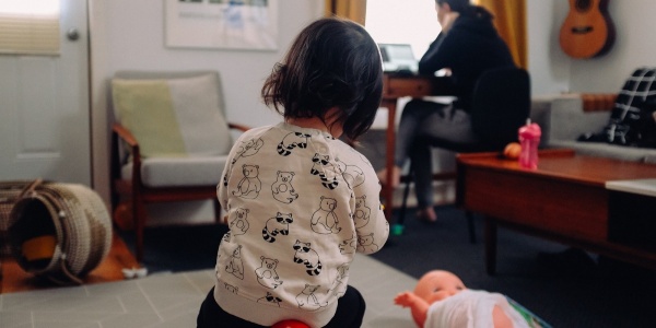 bimba in primo piano di spalle che gioca in casa e sullo sfondo la mamma che lavora al pc