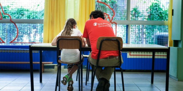 operatore save the children di spalle seduto a un tavolo con affianco una bambina seduta