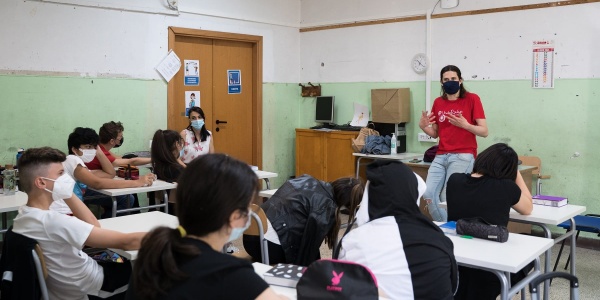 classe di studenti ripresi da dietra seduti ai banchi, in fondo un insegnante in piedi appoggiato alla cattedra spiega