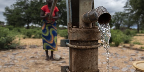 In primo piano una fontana a pompa da cui sgorga acqua, dietro sfocata si vede una donna che aziona la pompa per far uscire l acqua