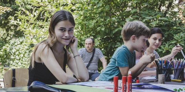 adolescente ucraina seduta a un banco insieme a un bambino biondo a fianco lei, girata verso la camera e appoggiata con il viso alla mano