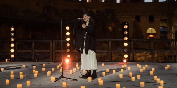 La cantautrice Elisa canta in piedi con microfono all interno del Colosseo circondata da candele accese sul palco. Elisa indossa un vestito lungo bianco e una giacca lunga nera