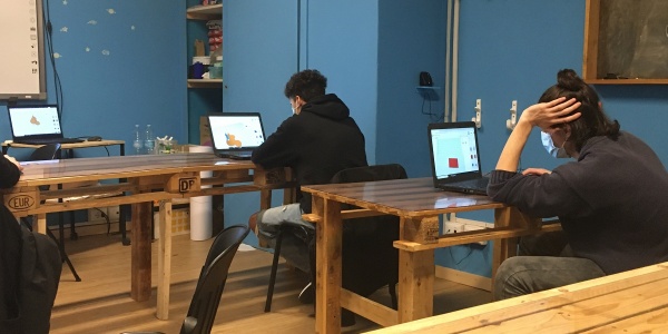 Stanza con tavoli di legno a cui sono seduti due ragazzi adolescenti al computer e ripresi di spalle. Indossano entrambi felpe di colore scuro.