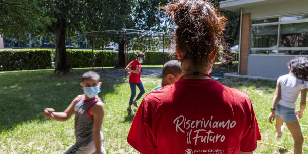 ragazza di spalle con maglietta rossa insieme a dei bambini che giocano nel prato con indosso la mascherina