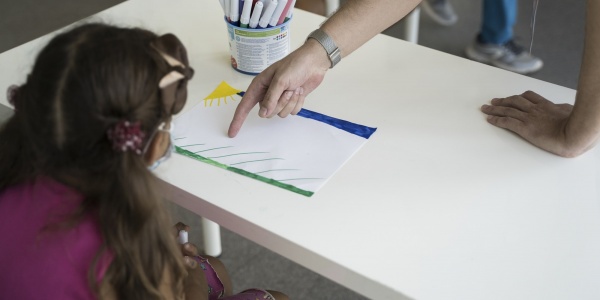 Bambina con codini seduta presa di spalle davanti a un tavolo bianco e davanti a lei c è il braccio di un adulta che indica un foglio sul tavolo