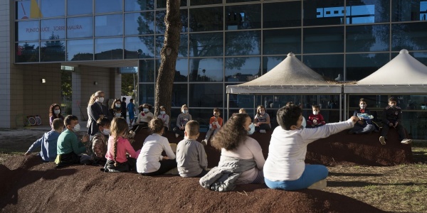 Foto all aperto, una fila di bambini di spalle seduti su un terrapieno guardano due gazebo bianchi montati di fronte a loro