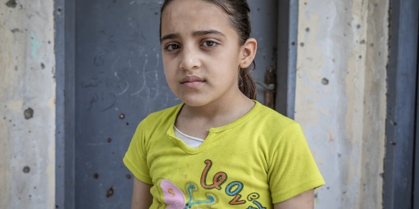 Mezzo busto di una bambina palestinese che indossa una maglietta gialla a maniche corte con alcune scritte colorate sopra. La bambina è castana ed ha i capelli raccolti in una coda.