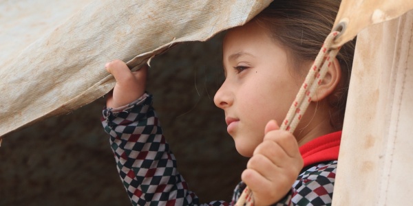 bambina siriana guarda verso sinistra attraverso una tenda che tiene aperta con una mano
