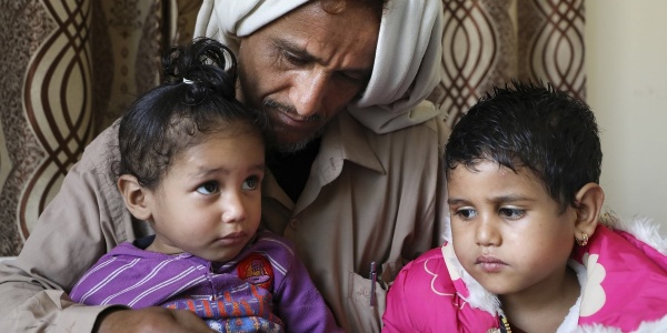 Un papà yemenita con turbante bianco in testa tiene in braccio le sue due bambine e le guarda. Entrambe hanno i capelli corti e neri, una guarda verso l alto e l altra verso la sua sorellina. Indossano magliette viola e rosa