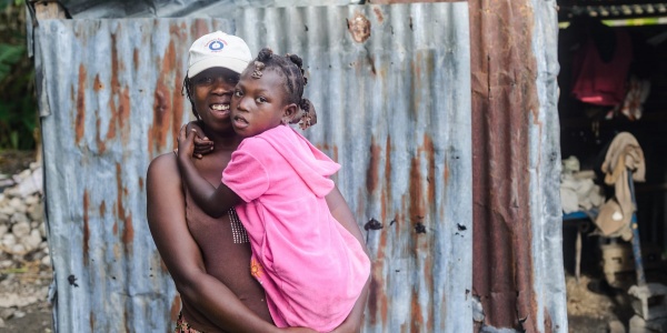 Una donna haitiana tiene in braccio la sua bambina che indossa un vestito rosa e le tiene le braccia strette intorno al collo. La donna indossa una maglietta marrone e un cappellino bianco in testa. Dietro di loro si vedono le lamiere di una baracca.