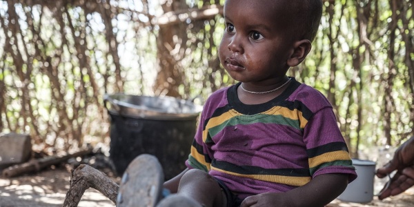 Bambino africano seduto a terra riparato in una capanna indossa una maglietta viola a strisce gialle e guarda alla sua destra.