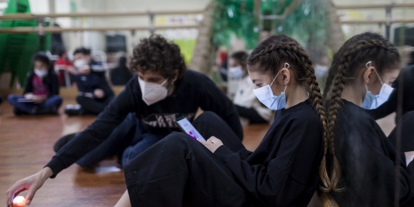 una ragazza e un ragazzo vestiti in tuta nera seduti per terra in un aula di danza, indossano mascherina chirurgica per covid-19, lei legge lui gioca con alcuni giochini per terra