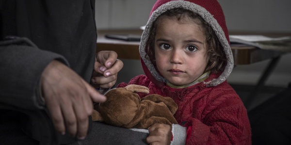 primo piano bambina siriana con cappotto e cappuccio rosso in testa stringe orsacchiotto appoggiata alla gamba di un adulto