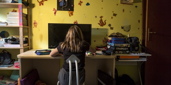 ragazza di spalle seduta alla scrivania della sua cameretta davanti al pc. Intorno a lei giochi da tavola, libri e quaderni.