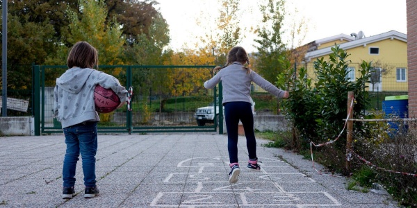 una bambina sulla sinistra tiene un pallone sotto il braccio mentre guarda l'amica che salta sui riquadri a terra del gioco campana