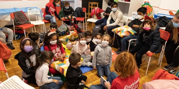 bambini e bambine seduti a terra in cerchio con mascherine che leggono un libro insieme all'operatrice
