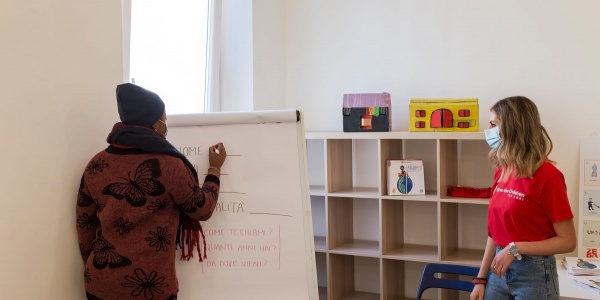 Una ragazza di spalle scrive su una lavagna e un operatrice di Save the Children la guarda all interno del nuovo CivicoZero di Torino