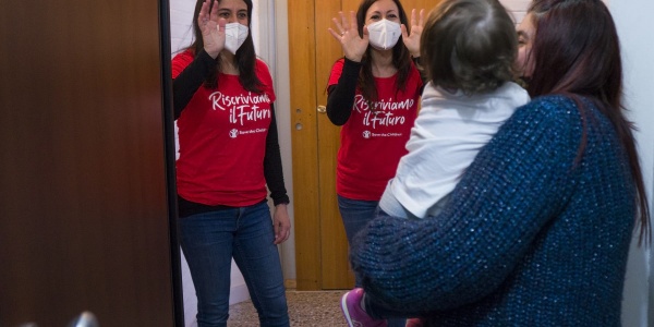 Due operatrici di Save the Children con maglietta della campagna Riscriviamo il Futuro salutano mamma e figlia davanti a loro sull uscio della porta di casa