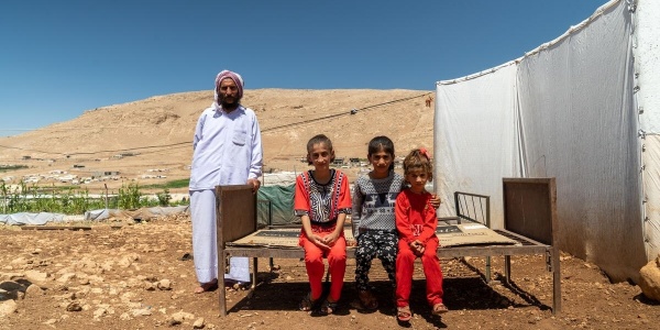 tre bambini yazidi seduti su una panca e in piedi alla loro destra il padre vestito di bianco 