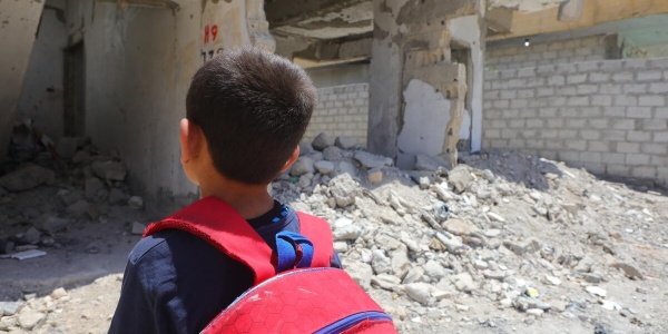 bambino con zaino rosso che guarda le macerie di una scuola distrutta