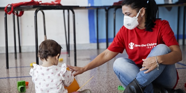 operatrice save the children con maglietta rossa con logo seduta per terra con una bimba di spalle mentre giocano insieme