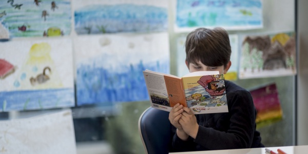 bambino seduto su una sedia davanti a un banco di scuola mentre legge un libro che tiene di fronte al suo viso