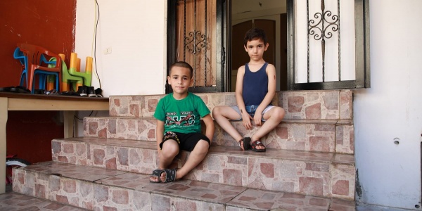 due bambini sono seduti sugli scalini di una casa, indossano abiti estivi. Uno ha la maglietta verde l altro blu