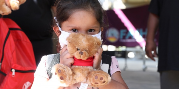 bimba con capelli castano scuro, bianca di carnagione, indossa una mascherina per proteggersi dal covid-19 e tiene tra le mani di fronte al viso un peluche a forma di orsacchiotto