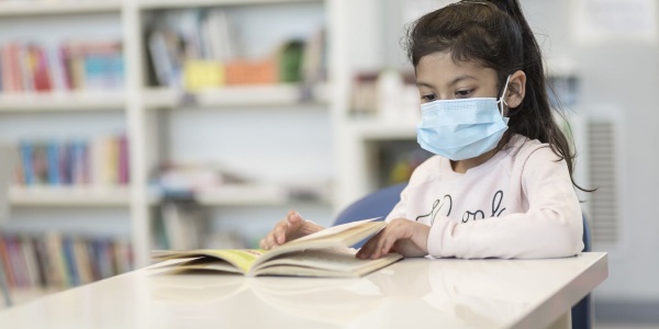 Bambina mora con coda ai lunghi capelli indossa mascherina chirurgica sul volto e una maglietta rosata legge un libro seduta a un tavolino