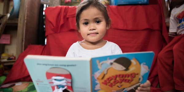 bimba del sud est asiatico con occhi scuri e capelli castani seduta con un libro grande quanto lei aperto innanzi a sè. 