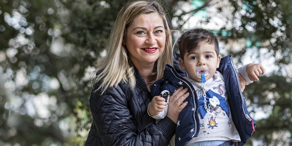 Una mamma bionda con capelli fino alle spalle è molto sorridente e ha occhi chiari, mantiene in piedi suo figlio di circa un anno con il ciuccio in bocca.