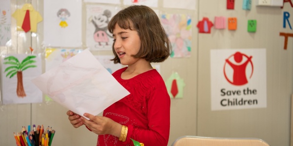 bambina in classe, in piedi, mentre legge ad alta voce qualcosa da un foglio. Sullo sfondo la parete dell aula è ghermita di disegni dei bambini e delle bambine.