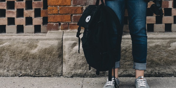 gambe di una studentessa che tiene in mano uno zaino nero. Sullo sfondo muro di mattoncini