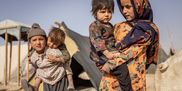 Quattro bambini afgani: una bambina tiene in braccio un bambino più piccolo, un altro bambino tiene sulle spalle un bimbo piccolo. Sullo sfondo le tende del loro villaggio